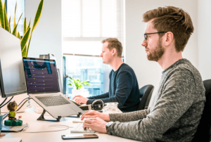 Two men sit side by side at a desk, working on computers in a bright office space. One uses a laptop on a stand with code on the screen; the other uses a desktop monitor. A potted plant is on the desk near the window. | Humanico Workforce Intelligence Software