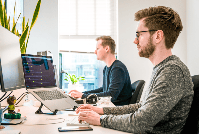 Two men sit side by side at a desk, working on computers in a bright office space. One uses a laptop on a stand with code on the screen; the other uses a desktop monitor. A potted plant is on the desk near the window. | Humanico Workforce Intelligence Software