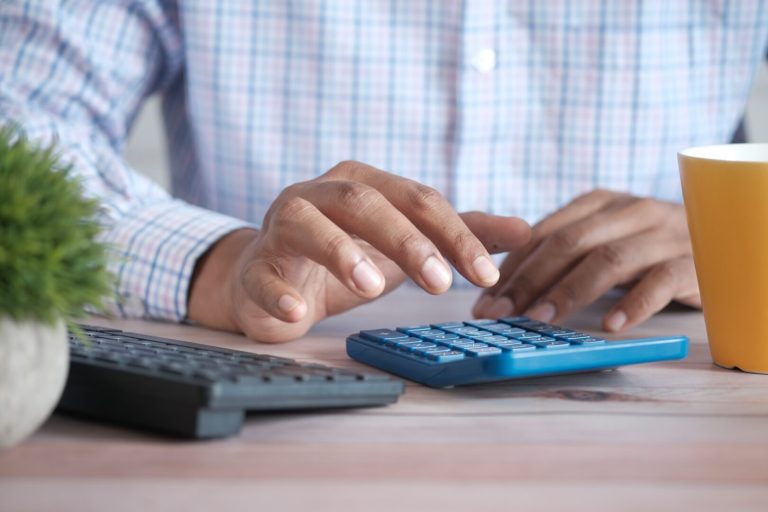 A person wearing a plaid shirt is using a blue calculator on a wooden desk, surrounded by HR technology tools—a black keyboard, yellow mug, and small potted plant nearby. | Humanico Workforce Intelligence Software