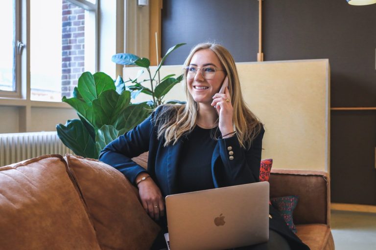 A woman with long blond hair, wearing glasses and a dark blazer, sits on a sofa with a laptop on her lap. She is smiling and talking on her phone in a bright, modern HR office setting with plants in the background. | Humanico Workforce Intelligence Software