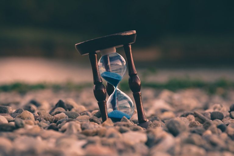 A wooden hourglass with blue sand stands tilted on a bed of small, smooth stones outdoors, capturing milestone moments amid blurred greenery and soft light in the background. | Humanico Workforce Intelligence Software