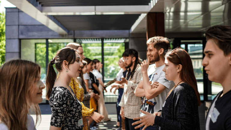 A group of young adults stands in two facing lines, engaging in lively conversations about post merger integration under a covered outdoor area with large windows and natural light. | Humanico Workforce Intelligence Software