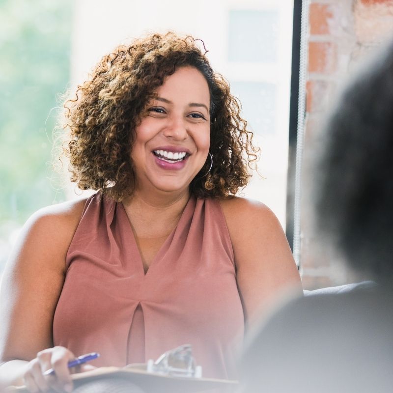 A woman with curly hair wearing a sleeveless pink top smiles warmly while holding a pen and clipboard, sitting indoors near a window—ready to discuss team engagement. | Humanico Workforce Intelligence Software
