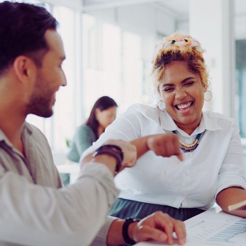 Two colleagues smiling and giving each other a fist bump at a desk in a bright office, with another person working in the background. The friendly and positive atmosphere reflects strong employee retention. | Humanico Workforce Intelligence Software