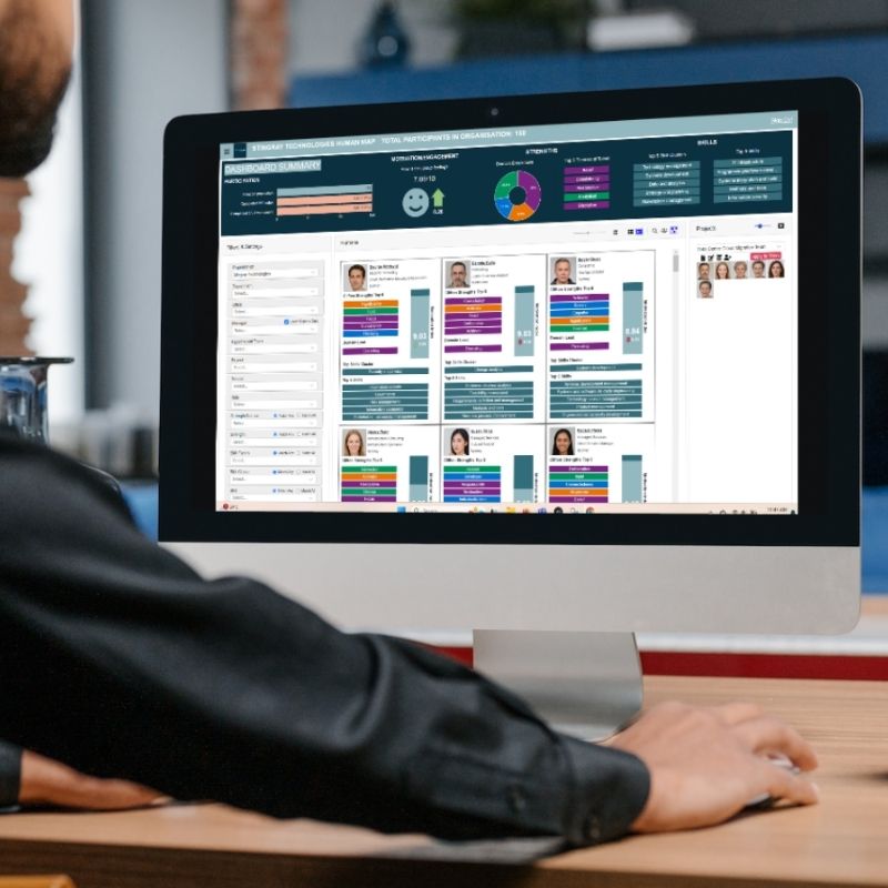 A person sitting at a desk uses a desktop computer displaying colorful data charts, graphs, and profile images on a dashboard interface powered by workforce intelligence software for tracking team engagement and performance. | Humanico Workforce Intelligence Software