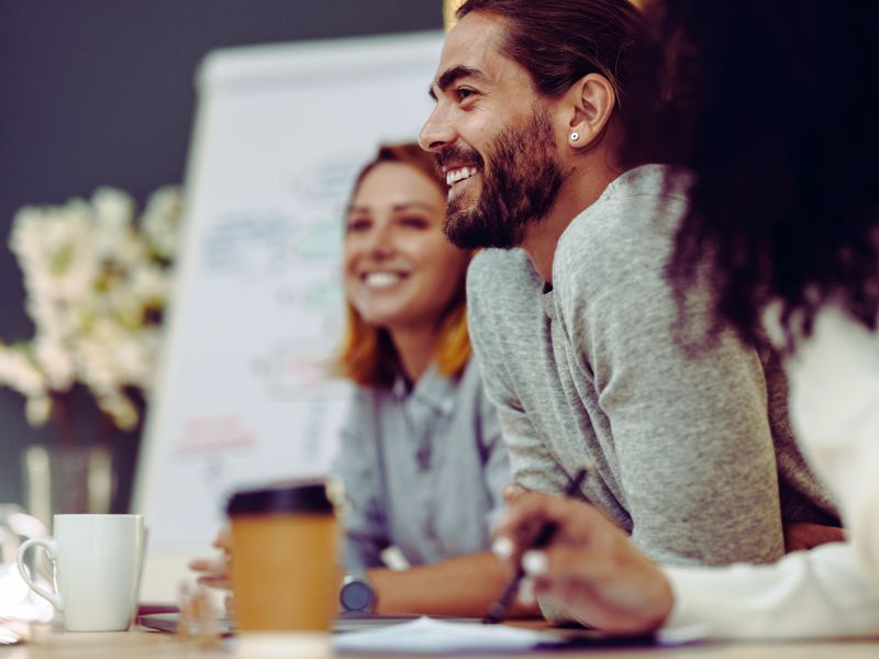 Two people sit at a table, smiling and engaged in conversation. A man with a beard and long hair is in focus, while a workforce intelligence platform helps boost team performance, visible in notes on the whiteboard behind them. | Humanico Workforce Intelligence Software