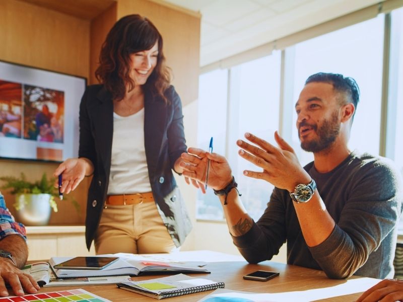 Two colleagues, a woman standing and a man sitting, engage in a lively discussion at a sunlit office table. Smiling and animated, they collaborate over notebooks and papers—just another inspiring moment as you meet the Humanico Team. | Humanico Workforce Intelligence Software