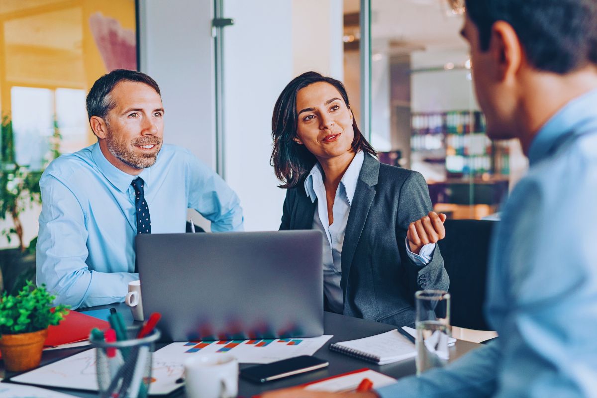 Two professionals in business attire sit at a table with a laptop and documents, discussing skills mapping with a third person across from them in a modern office setting. | Humanico Workforce Intelligence Software