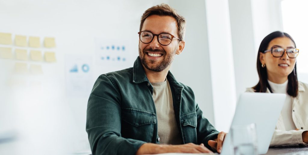 Two people sit at a desk in a bright office, smiling and looking off-camera. They appear engaged in a discussion about hr tools for executives, with one man in glasses and a green jacket and a woman with glasses and long dark hair. | Humanico Workforce Intelligence Software