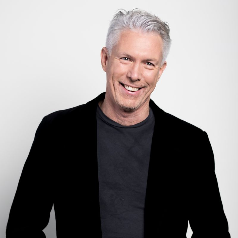 Smiling man with short gray hair, a black jacket over a dark shirt, stands against a plain white background—perfect for hr tech founders or workforce intelligence founders seeking a professional image. | Humanico Workforce Intelligence Software