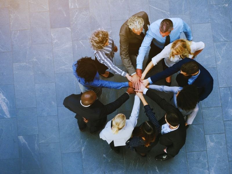 A group of business people stands in a circle, stacking their hands together to symbolize teamwork and unity—contact Humanico for people analytics consultation to strengthen your team. | Humanico Workforce Intelligence Software