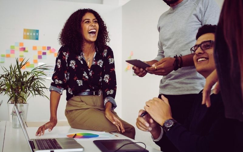 A woman laughs while sitting on a desk with colleagues gathered around her, holding devices and smiling—a collaborative atmosphere that helps develop leaders and reduce staff turnover. Colorful sticky notes cover the wall, and a potted plant sits on the desk. | Humanico Workforce Intelligence Software