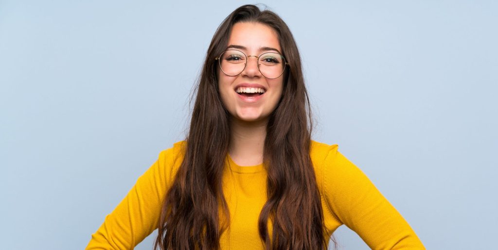 A young woman with long brown hair and glasses smiles while standing against a plain light blue background. She is wearing a bright yellow long-sleeve shirt, ready to discuss onboarding for new managers or explore the latest HR tools. | Humanico Workforce Intelligence Software