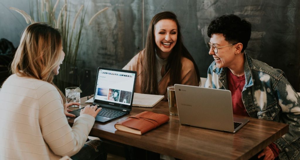 Three people sit at a wooden table with laptops, notebooks, and drinks, smiling and laughing together in a casual, modern setting—capturing the spirit of workforce intelligence founders behind Humanico. | Humanico Workforce Intelligence Software