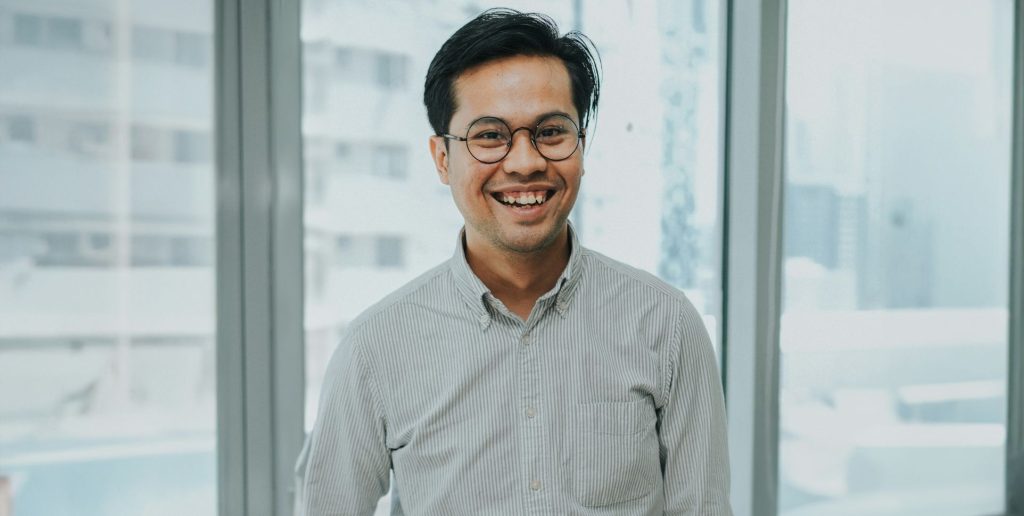 A man wearing glasses and a striped shirt smiles while standing indoors in front of large windows with an urban background, representing the positive impact of HR tools for executives. | Humanico Workforce Intelligence Software
