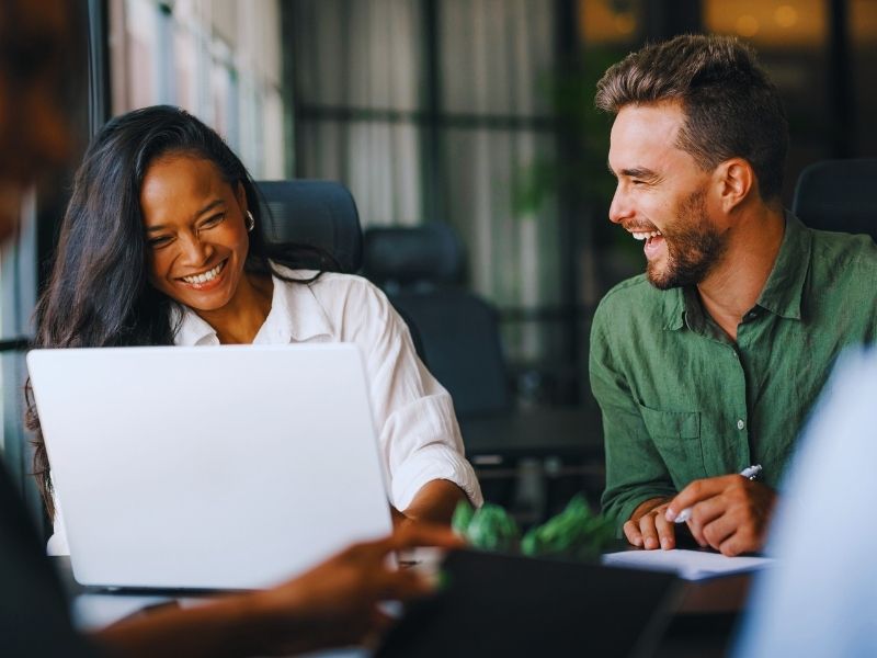 A woman and a man sit at a table in an office, smiling and laughing together. The woman works on a laptop during their people analytics consultation, while the man in a green shirt looks at her. The atmosphere is relaxed and friendly—contact Humanico to learn more. | Humanico Workforce Intelligence Software