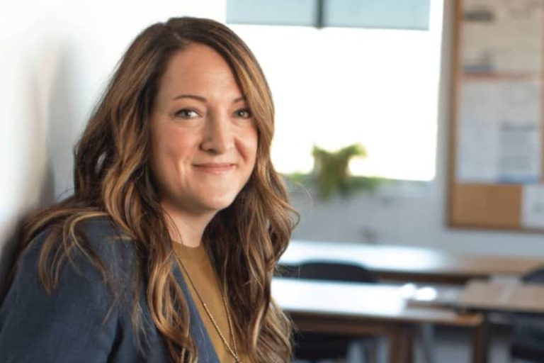 A woman with long wavy brown hair, wearing a blue blazer and mustard shirt, smiles while sitting in a bright, modern classroom—an inspiring setting to discuss Humanico’s powerful HR software and workforce intelligence platform. | Humanico Workforce Intelligence Software