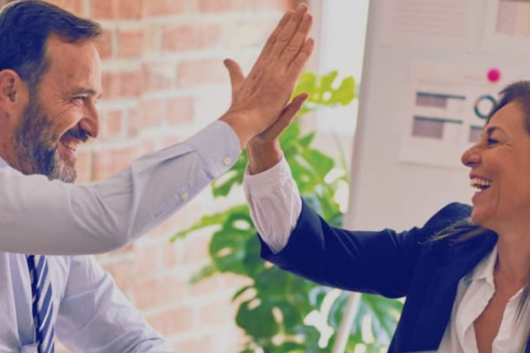 Two smiling people in business attire give each other a high-five in an office setting, celebrating success with the help of workforce intelligence platform humanico. A plant and a brick wall are visible in the background. | Humanico Workforce Intelligence Software
