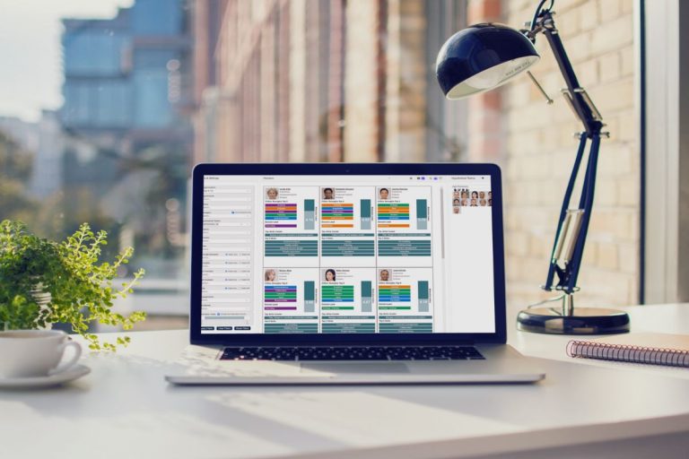 A laptop on a desk displays a grid of colorful profile cards, showcasing the power of Humanico HR software. A lamp, notepad, and potted plant sit nearby, while large windows reveal a modern cityscape in the background. | Humanico Workforce Intelligence Software