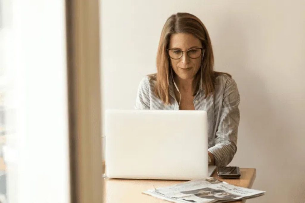 A woman with glasses and shoulder-length hair sits at a desk, working on her laptop. A smartphone and newspapers rest beside her as she explores workforce intelligence platform options like Humanico. The background is plain and light-colored. | Humanico Workforce Intelligence Software