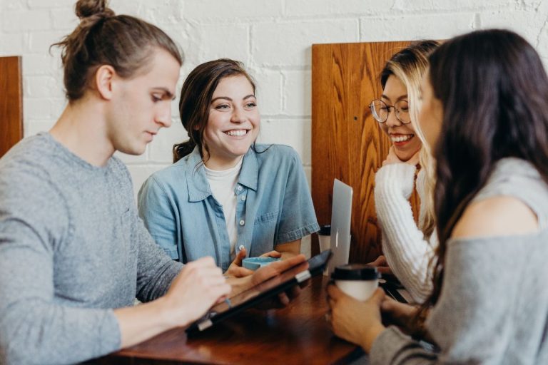 Four young adults sit together at a table, smiling and talking. One writes on a tablet—perhaps exploring HR software like Humanico—while another holds a coffee cup. They appear to be having a casual, friendly conversation in a cozy setting. | Humanico Workforce Intelligence Software
