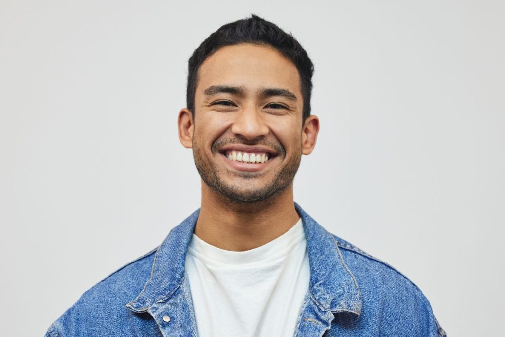 A young man with short dark hair and light facial hair smiles broadly. He is wearing a white t-shirt and a blue denim jacket, embodying the fresh energy of the modern HR tech landscape. The background is plain and light-colored. | Humanico Workforce Intelligence Software