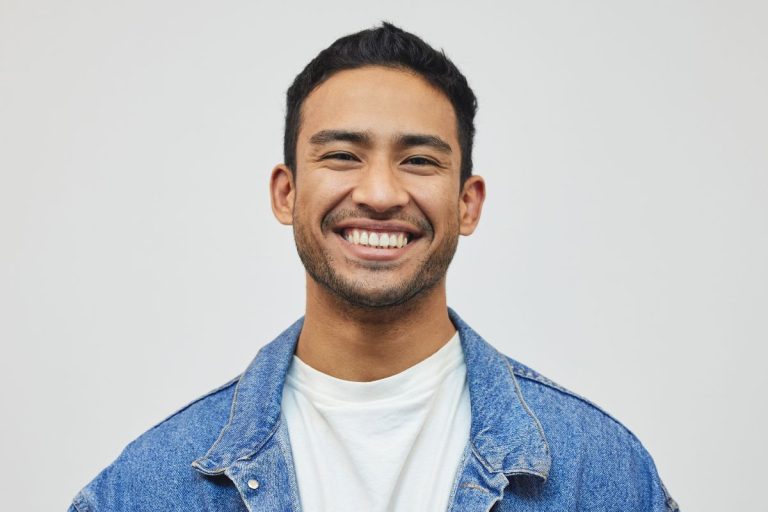 A young man with short dark hair and light facial hair smiles broadly. He is wearing a white t-shirt and a blue denim jacket, embodying the fresh energy of the modern HR tech landscape. The background is plain and light-colored. | Humanico Workforce Intelligence Software