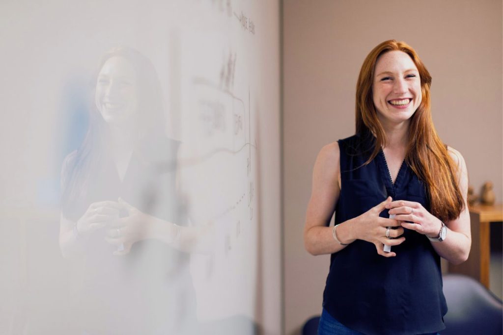 A woman with long red hair, wearing a navy sleeveless blouse, stands and smiles confidently in an office next to a whiteboard with drawings and notes on HR tech and employee retention. Her reflection is visible on the glass surface. | Humanico Workforce Intelligence Software