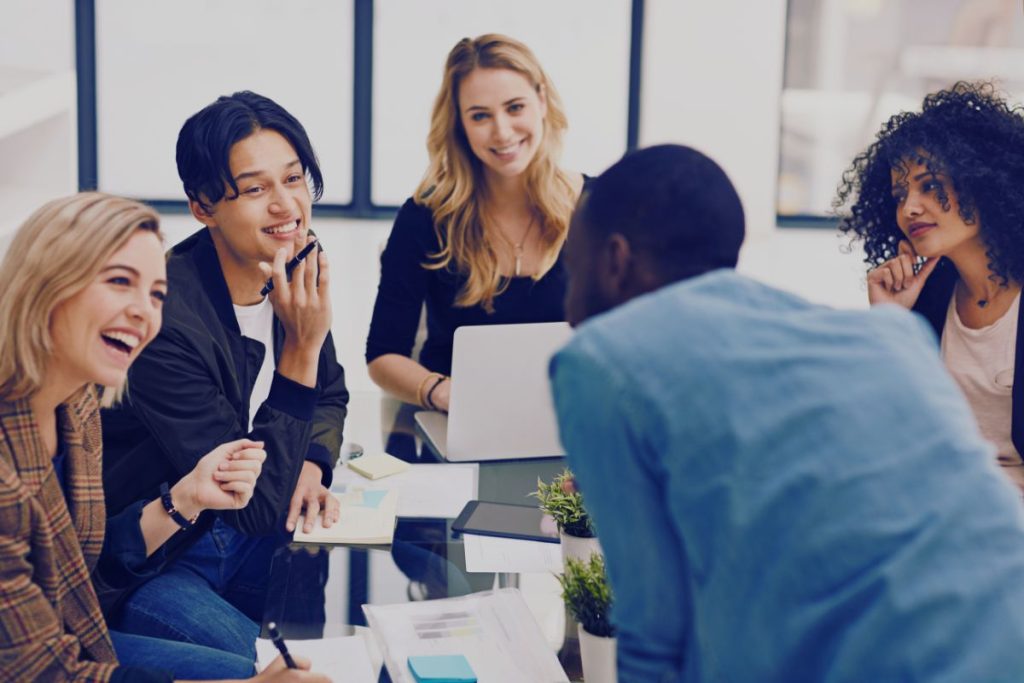 Five young adults sit around a glass table in an office, smiling and talking. Laptops, papers, and plants are on the table—an energizing Humanico brainstorming session focused on employee retention. Bright windows shine in the background. | Humanico Workforce Intelligence Software