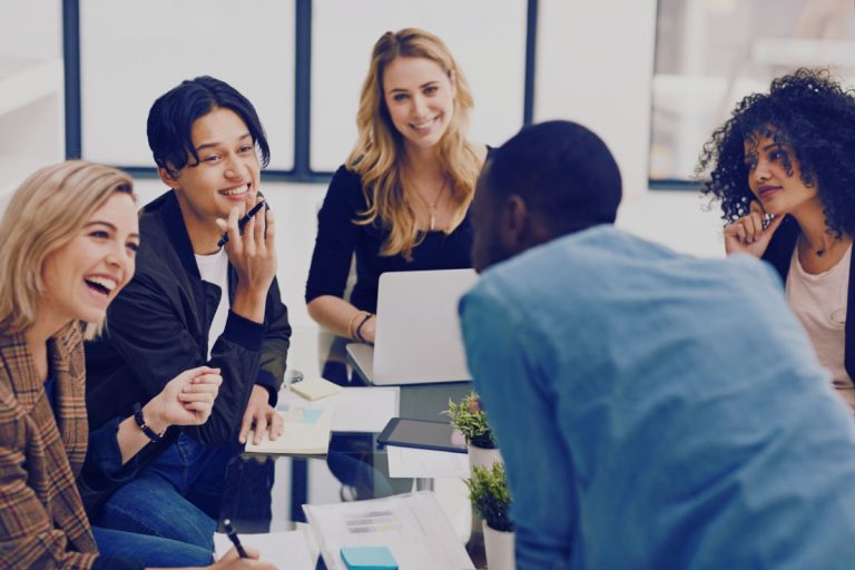 Five young adults sit around a glass table in an office, smiling and talking. Laptops, papers, and plants are on the table—an energizing Humanico brainstorming session focused on employee retention. Bright windows shine in the background. | Humanico Workforce Intelligence Software
