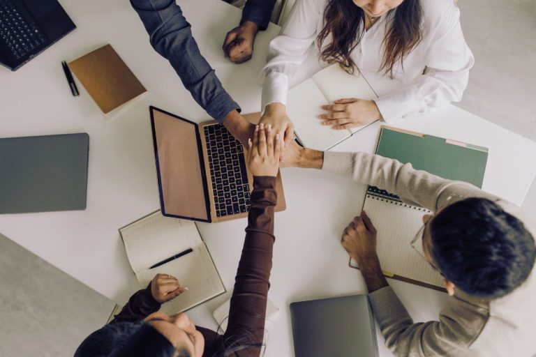 Five people sit around a table with notebooks, a laptop, and folders, placing their hands together in a gesture of teamwork and unity—demonstrating how Humanico HR software can boost employee retention through collaboration. | Humanico Workforce Intelligence Software