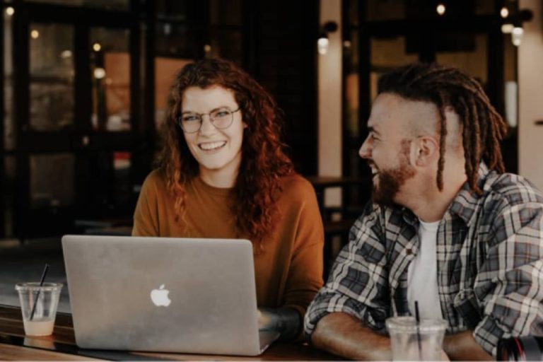Two people sit at a table outside, smiling and talking. One is working on a laptop with new HR software, while the other looks at them. Both have iced drinks. The background shows a building with large windows. | Humanico Workforce Intelligence Software