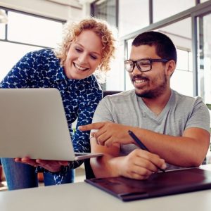 Two people smiling and looking at a laptop screen together in a bright office, collaborating on ways to improve team engagement. One person stands holding the laptop, while the other sits and points at the screen. | Humanico Workforce Intelligence Software