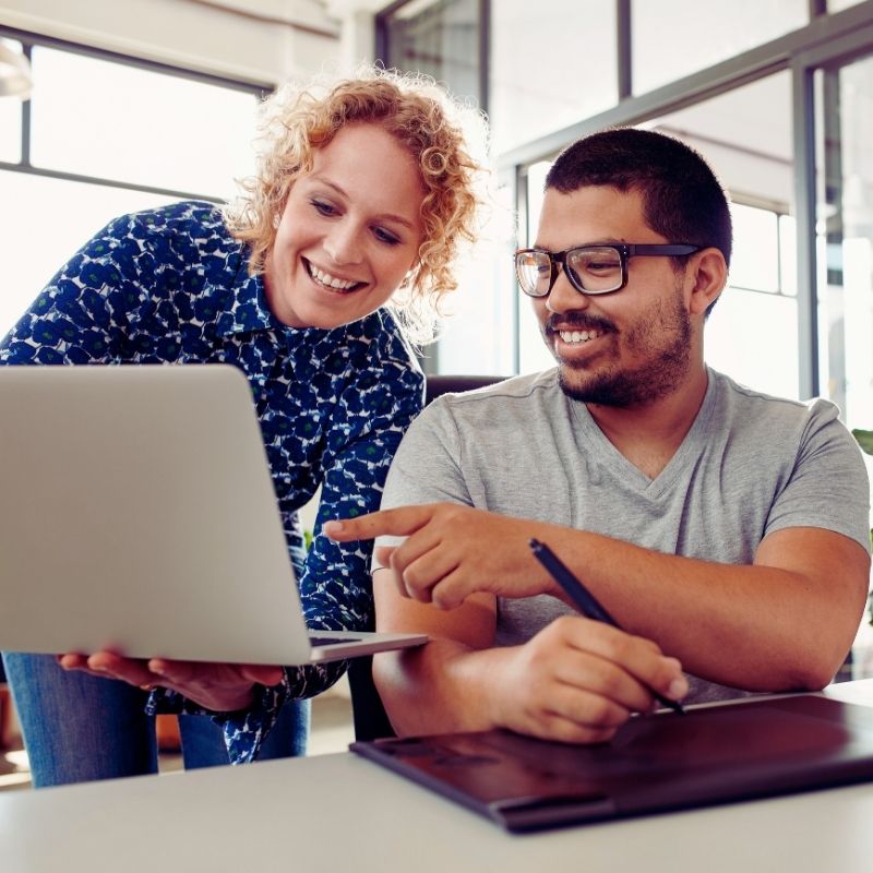 Two people smiling and looking at a laptop screen together in a bright office, collaborating on ways to improve team engagement. One person stands holding the laptop, while the other sits and points at the screen. | Humanico Workforce Intelligence Software