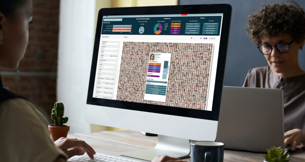Two people work at a desk with laptops. The main focus is a large computer screen displaying workforce intelligence founders’ dashboard with graphs, charts, and a grid of profile photos. A small plant is beside the monitor. | Humanico Workforce Intelligence Software