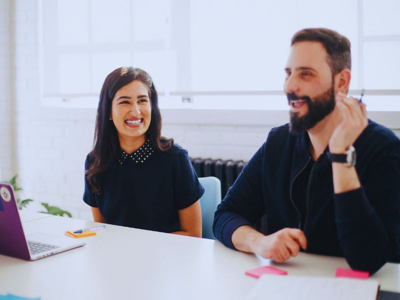 Two people sit at a table in a bright office, smiling and talking during a Humanico demo. One has long dark hair and wears a navy top; the other has a beard and wears a dark sweater. A laptop and notepads are on the table. | Humanico Workforce Intelligence Software