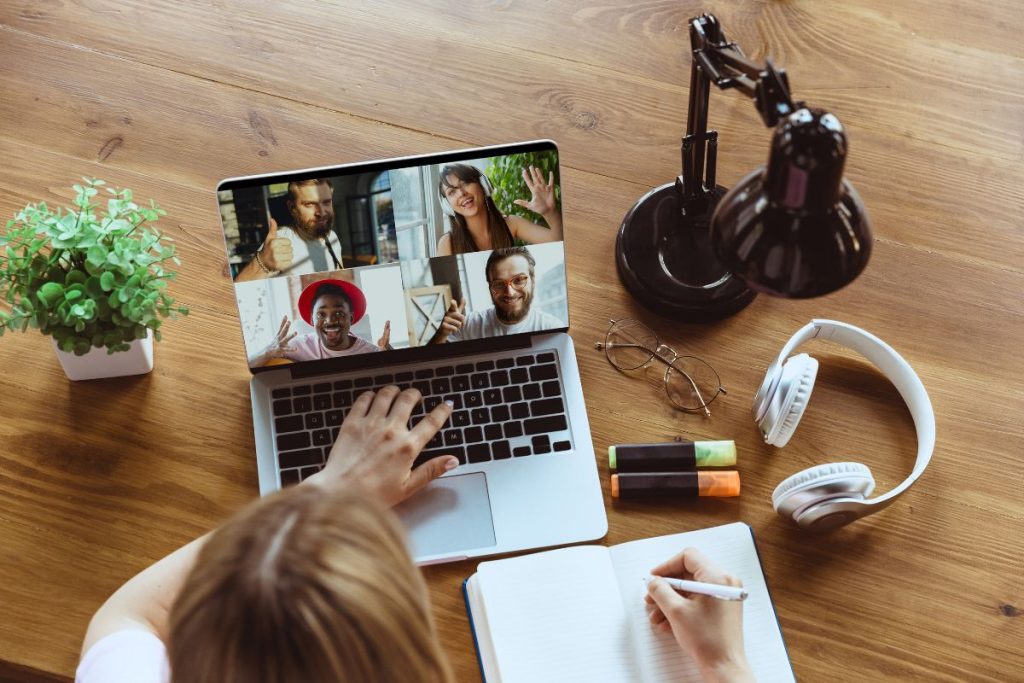 A person takes notes in a notebook while participating in an HR tech video call with four people on a laptop. The desk also has a lamp, plant, headphones, and colorful markers. | Humanico Workforce Intelligence Software
