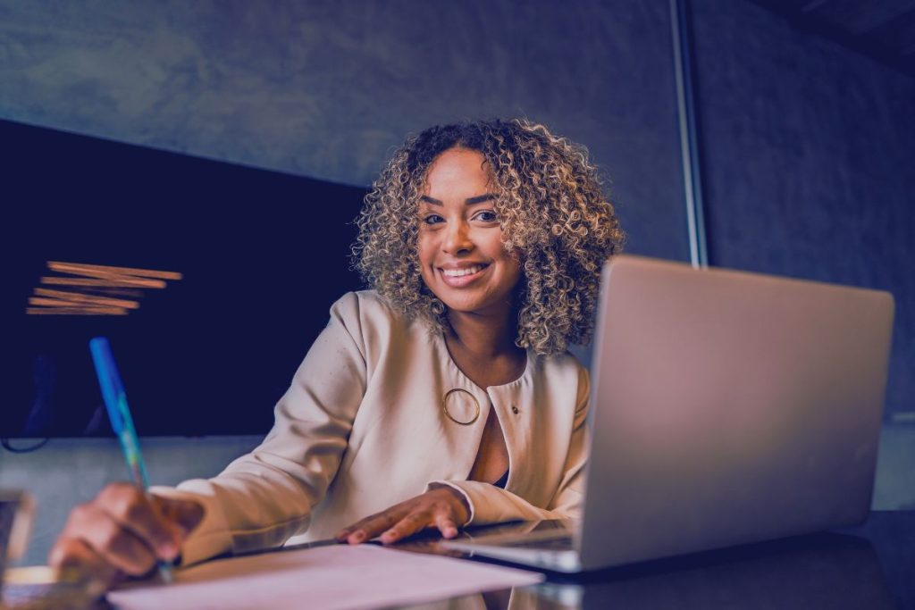 A woman with curly hair smiles while sitting at a desk with an open laptop, jotting notes on a document. She is wearing a light-colored blazer and appears to be working in an office, exploring HR tech solutions for employee retention. | Humanico Workforce Intelligence Software