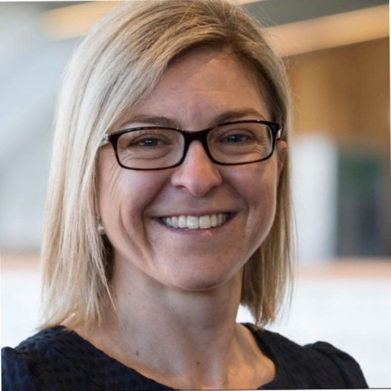A woman with straight blonde hair and black glasses smiles at the camera. She is wearing a dark top and is in an indoor setting with a softly blurred background. | Humanico Workforce Intelligence Software