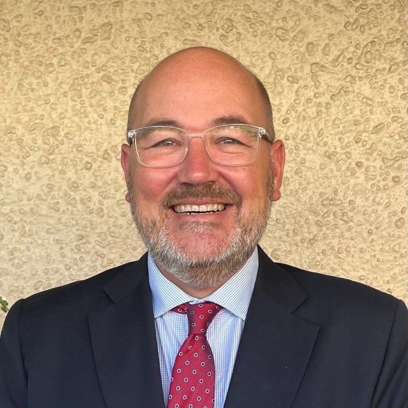 A smiling man with glasses, a mustache, and a beard wears a dark suit, light blue shirt, and red patterned tie, standing in front of a textured beige wall. | Humanico Workforce Intelligence Software