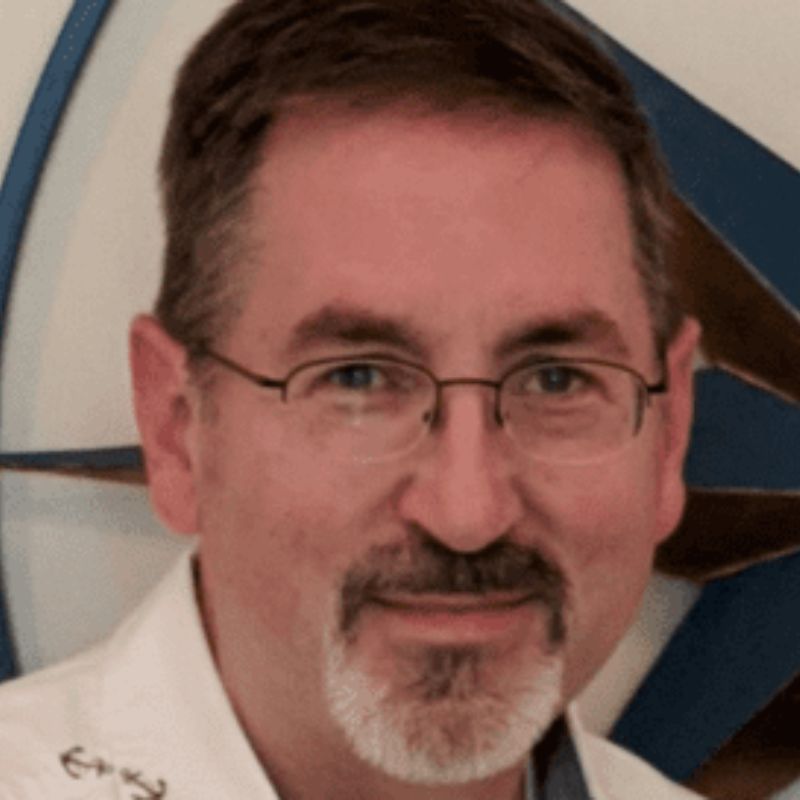 A man with short brown hair, glasses, and a goatee smiles at the camera. He is wearing a white shirt, and there is a blurred geometric design in blue and brown in the background. | Humanico Workforce Intelligence Software