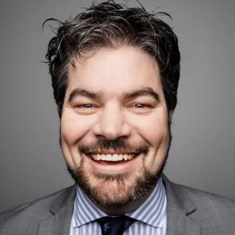 Tom Pennington, a man with dark, wavy hair and a beard, smiles broadly in a gray suit, blue-and-white striped shirt, and navy tie against a plain gray background. | Humanico Workforce Intelligence Software