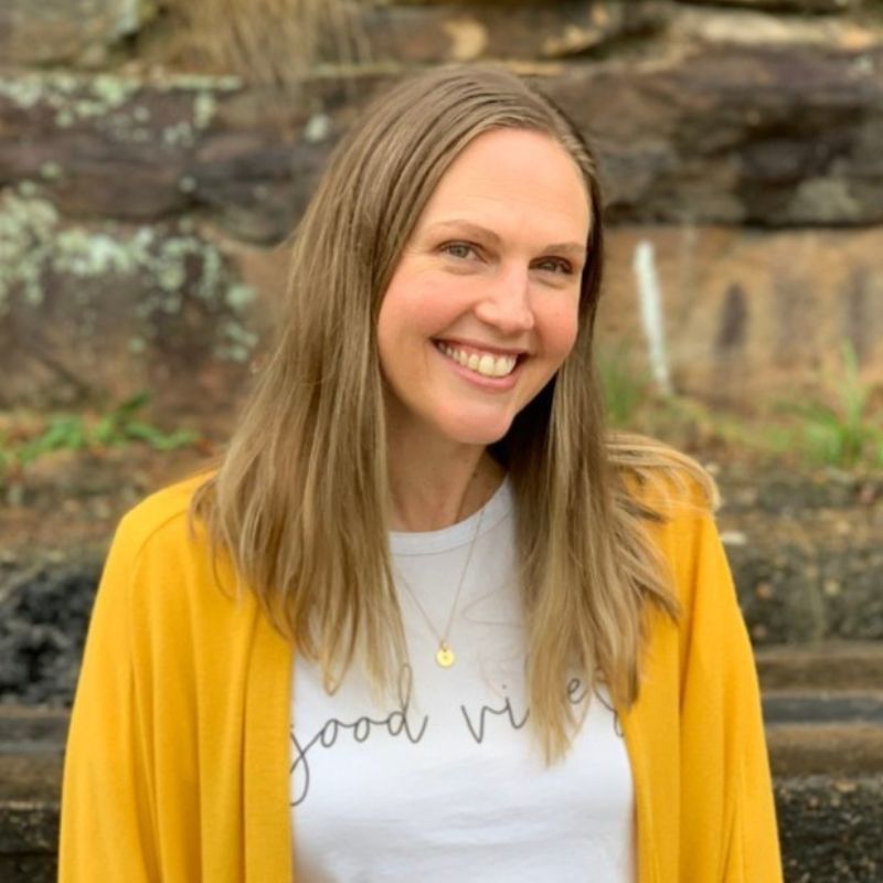 A woman with long, light brown hair smiles at the camera. She is wearing a white shirt with "good vibes" written on it and a mustard yellow cardigan. A stone wall and greenery are in the background, captured by Tom Pennington. | Humanico Workforce Intelligence Software