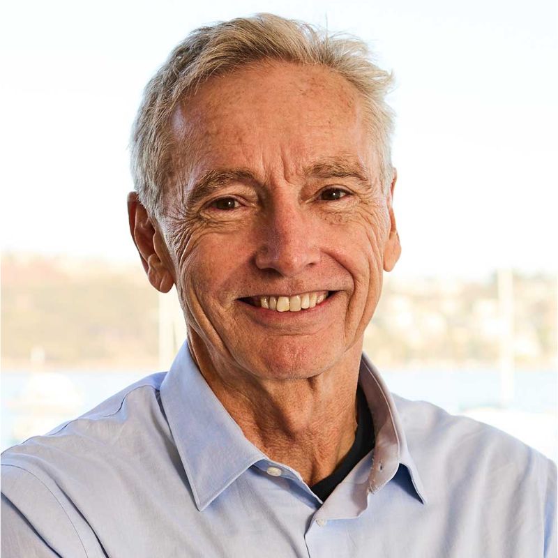 An older man with short gray hair smiles at the camera. He is wearing a light blue collared shirt. The background is softly blurred, showing a bright outdoor scene with water and hints of buildings. | Humanico Workforce Intelligence Software