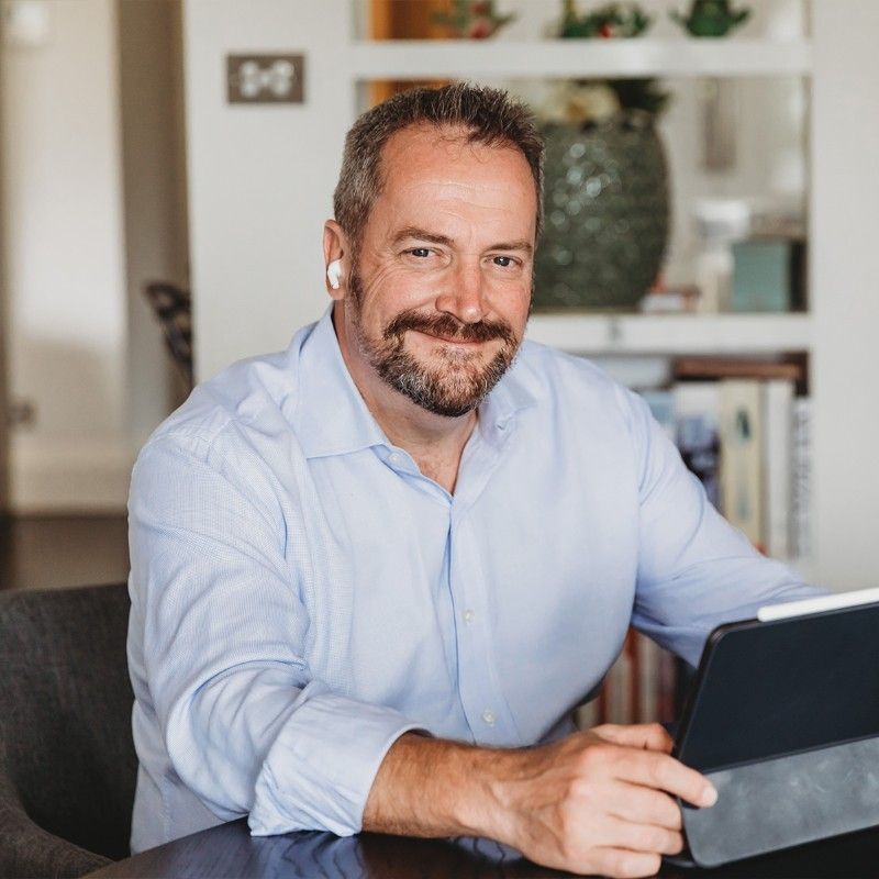 A man with short hair and a beard, wearing a light blue shirt and wireless earbuds, smiles while sitting at a table and using a tablet in a modern, well-lit room. | Humanico Workforce Intelligence Software