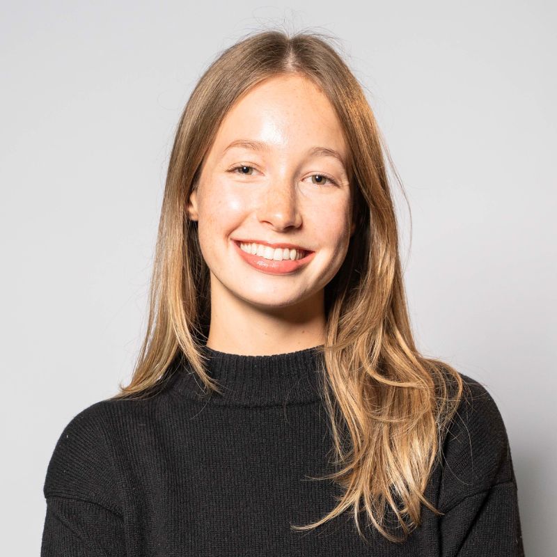 Scarlett Foulsham, a young woman with long, straight, light brown hair, smiles at the camera. She is wearing a black, long-sleeve sweater and stands against a plain, light gray background. | Humanico Workforce Intelligence Software