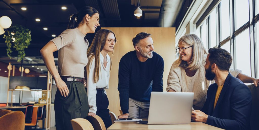 Five people gather around a table with a laptop, smiling and talking in a bright, modern office filled with large windows and indoor plants, as they discuss employee retention strategies using the latest HR software. | Humanico Workforce Intelligence Software