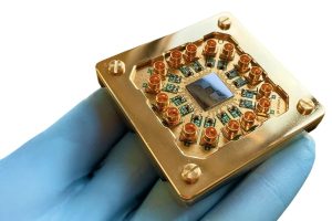 A gloved hand holds a gold-colored quantum computer chip with multiple connectors and a central black square component, highlighting innovation that goes beyond qualifications, set against a white background. | Humanico Workforce Intelligence Software