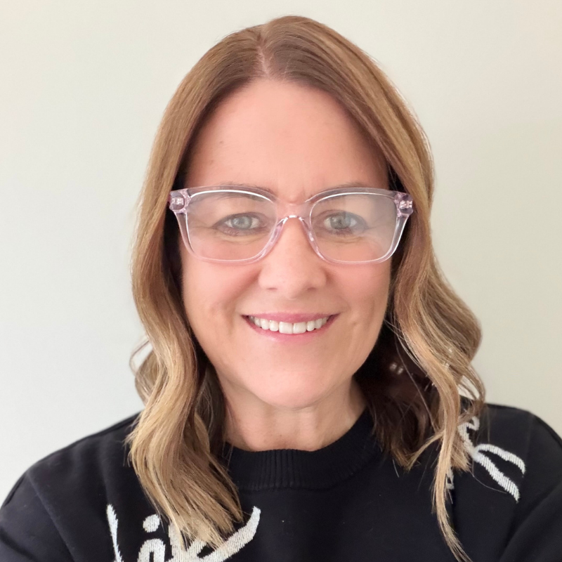 Julie Stuart, a woman with shoulder-length wavy light brown hair, wearing clear glasses and a black sweater with white writing, smiles in front of a plain light background. | Humanico Workforce Intelligence Software