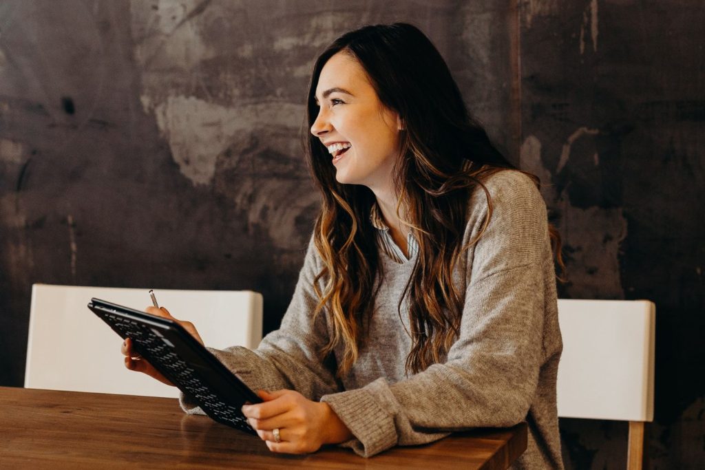 A woman with long brown hair, wearing a gray sweater, sits at a wooden table smiling while holding a tablet and stylus—discovering workforce wide insights—against a dark textured wall in the background. | Humanico Workforce Intelligence Software